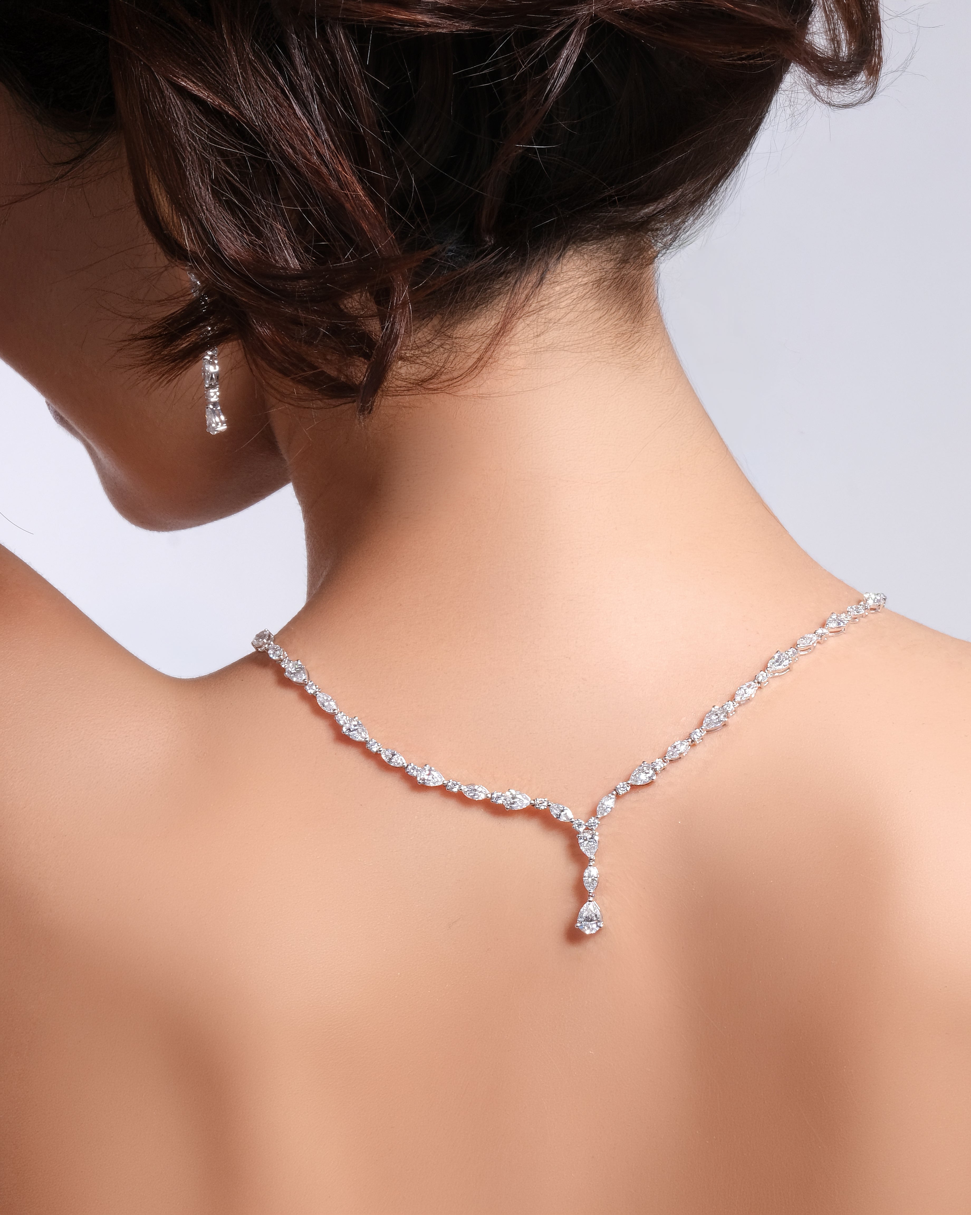 A close-up back view of a woman wearing a delicate Y-shaped diamond necklace and a matching drop earring. The jewelry is set in a silver-toned metal and features clear sparkling stones.